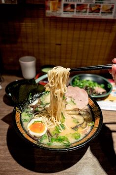 a person holding chopsticks over a bowl of noodles with broccoli and an egg