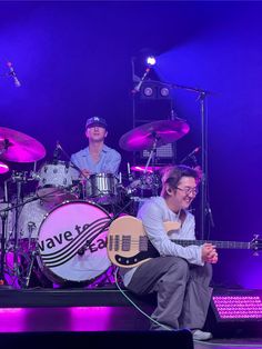 two men are playing instruments on stage with purple lighting behind them and one man is sitting in front of the drums