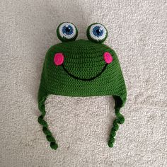 a green crocheted hat with eyes and a pink flower on the top, laying on a white surface