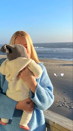 a woman holding a baby in her arms at the beach while wearing a knitted hat