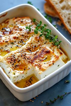 a white dish filled with cheese and herbs next to some slices of bread on a blue surface