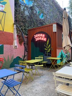 tables and chairs are outside in front of a colorful wall with an umbrella on it