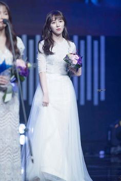 a woman in a white dress standing next to another woman wearing a white dress and holding flowers