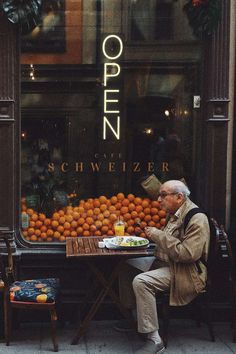 an old man sitting at a table with oranges in front of his store window