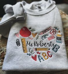 a white shirt with embroidered letters and school supplies in the shape of a heart that says mrs roberts's e - b c