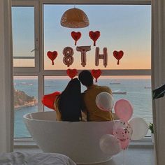 two people sitting in a bathtub with balloons on the window sill overlooking the ocean