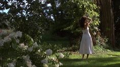 a woman in a white dress standing next to some trees and flowers on the grass