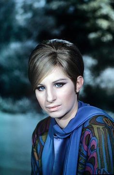 a woman with short hair wearing a blue scarf and looking at the camera while standing in front of trees