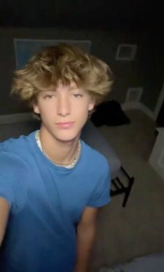 a young man with blonde hair wearing a blue t - shirt and silver necklace looking at the camera