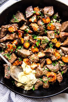 a skillet filled with meat and vegetables on top of a white table cloth next to a wooden spoon