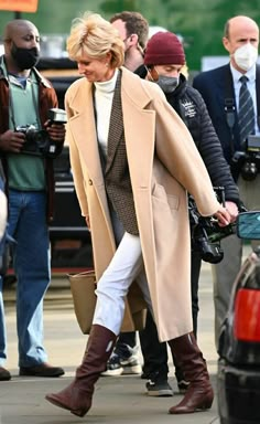 a woman walking down the street in white pants and brown boots, wearing a tan coat