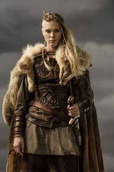 a woman dressed in medieval clothing with a fur collar and long hair standing on a cloudy day