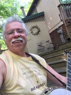 an older man with glasses and a beard holding a guitar in front of a house