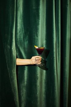 a woman holding a martini glass in front of a green curtain with her hand on it