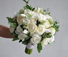 a bridal bouquet with white roses and greenery is held by someone's hand