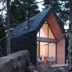 a small black cabin in the woods with chairs and tables outside at night, lit up by windows