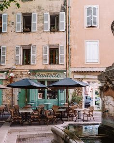 an outdoor cafe with tables and umbrellas next to a fountain in front of a building