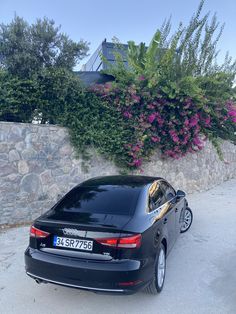 a black car parked in front of a stone wall with purple flowers growing on it