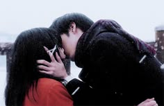 two people kissing in the snow with one person wearing a red jacket and black coat