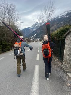 two people walking down the road with their skis strapped to their backs and backpacks on