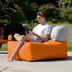 a young man sitting on an orange couch reading a book in the backyard with his legs crossed