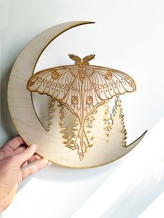 a hand holding a wooden cutout of a moth on a crescent with pine trees in the background