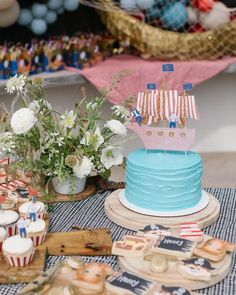 a blue cake topped with a pink sailboat on top of a wooden table next to cupcakes