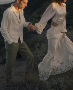 a man and woman are holding hands while walking through the grass in front of a hill