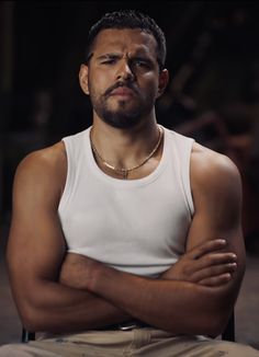 a man with his arms crossed sitting in a chair looking at the camera while wearing a white tank top