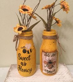 two mason jars with flowers in them sitting on a white tableclothed surface, one is decorated with bees and the other has words sweet as honey