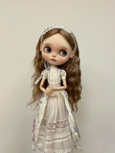 a doll with long hair wearing a white dress and headband, standing against a wall