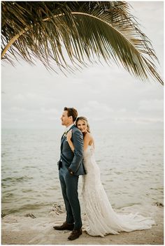 a bride and groom standing under a palm tree on the beach in front of the ocean