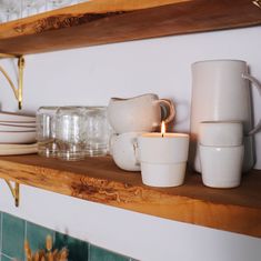 a shelf filled with dishes and cups on top of a tiled wall next to a candle