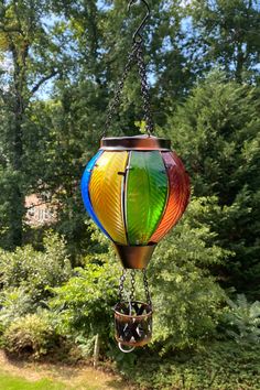 a multicolored glass lantern hanging from a chain in front of trees and bushes