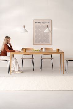 a woman sitting at a wooden table in front of a white wall with a poster on it