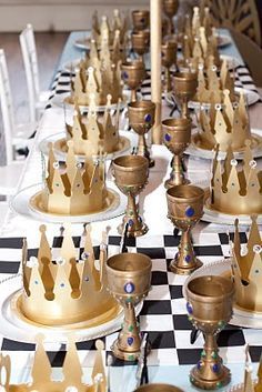 a table topped with lots of gold cups and plates covered in crowns on top of checkered cloth