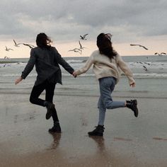 two people running on the beach with birds flying in the sky behind them and one person holding hands