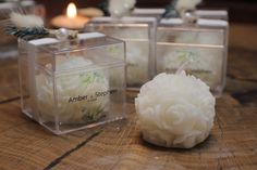 three clear boxes with white flowers in them on a wooden table next to candles and other decorations