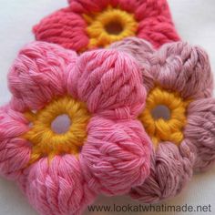 three crocheted flowers are sitting on a white tablecloth, one is pink and the other is yellow