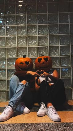 two people sitting on the ground with pumpkin heads in front of glass block wall behind them