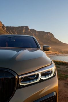 the front end of a silver car parked in front of a body of water with mountains in the background