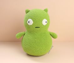 a green crocheted stuffed animal with eyes and ears sitting in front of a pink wall