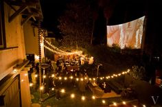 an outdoor movie screen is lit up at night with string lights strung across the lawn