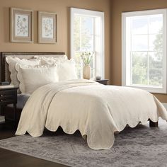 a bed with white comforter and pillows in a bedroom next to two large windows