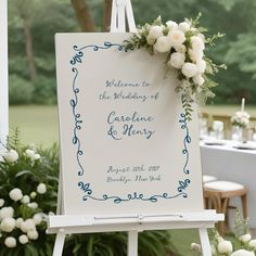 a welcome sign with white flowers on it and greenery in the background at a wedding