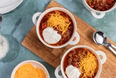 three white bowls filled with chili and cheese on top of a wooden cutting board next to spoons