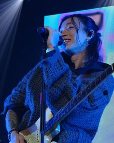 a woman holding a guitar and singing into a microphone while standing in front of a screen