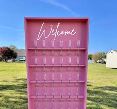 a pink welcome sign with wine glasses on it in front of a grassy field and house