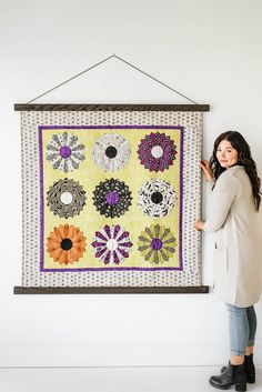 a woman standing next to a wall hanging on a white wall with a colorful quilt