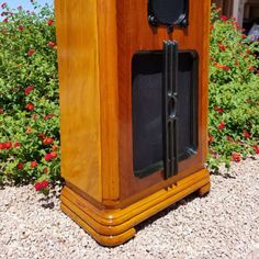 an old fashioned wooden clock sitting on top of a gravel ground next to red flowers